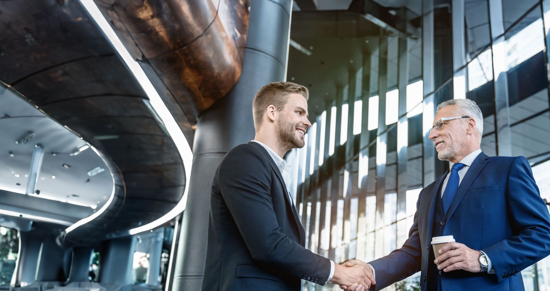 Two businessmen in formal suits shaking hands in a modern glass office building, symbolizing partnership, trust.